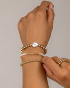Hand wearing multiple gold and pearl bracelets on a neutral background